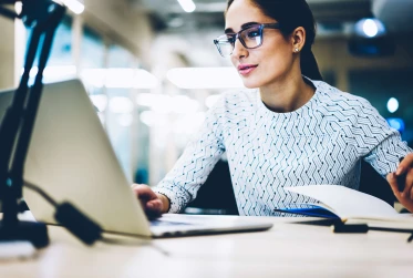 woman with glasses looking at laptop with pen in hand