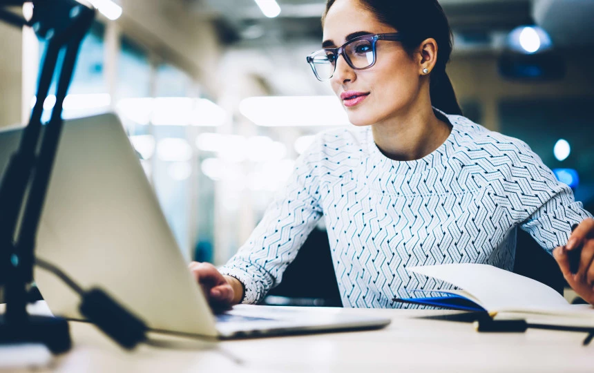 woman with glasses looking at laptop with pen in hand
