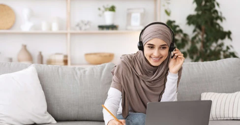 woman sitting on sofa wearing hijab and casual clothes working from home with headset and laptop