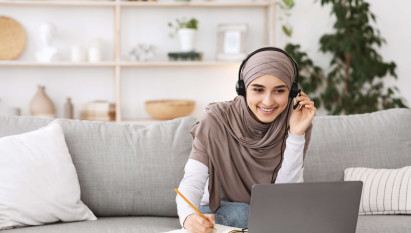 woman sitting on sofa wearing hijab and casual clothes working from home with headset and laptop