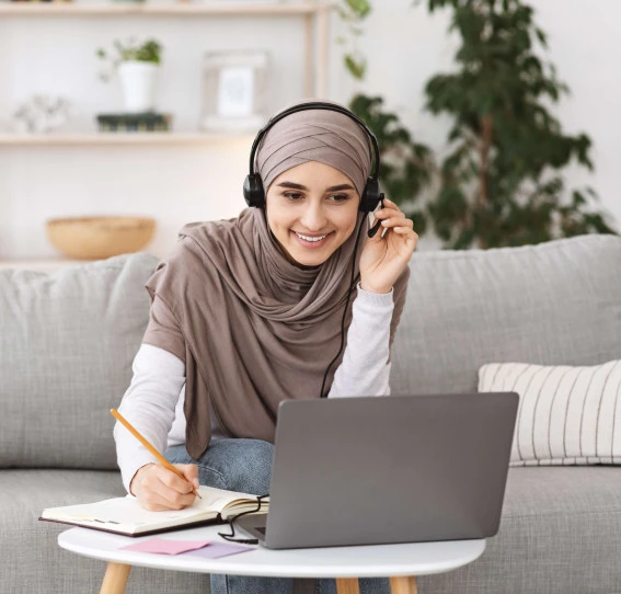 woman sitting on sofa wearing hijab and casual clothes working from home with headset and laptop