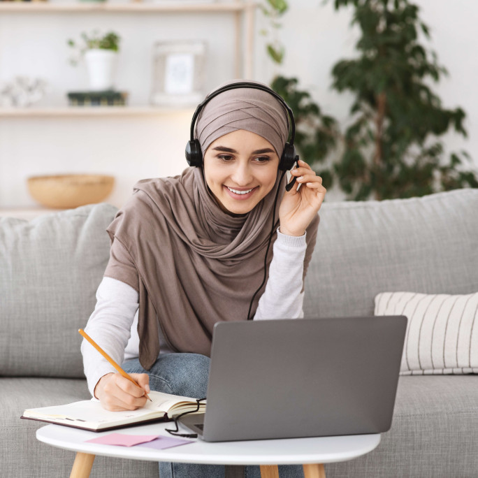 woman sitting on sofa wearing hijab and casual clothes working from home with headset and laptop