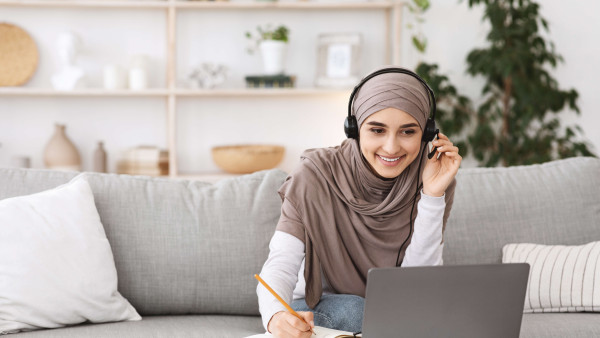 woman sitting on sofa wearing hijab and casual clothes working from home with headset and laptop