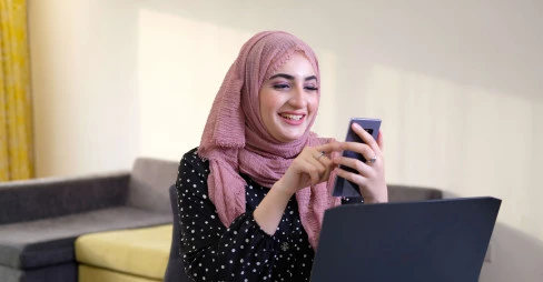 woman wearing hijab smiling at phone with laptop on table