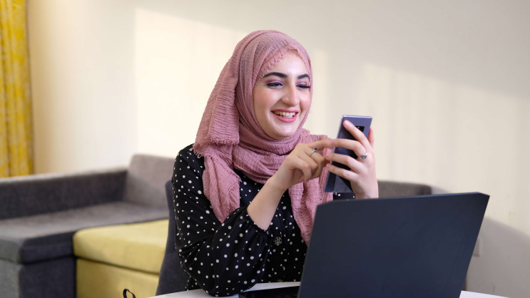 woman wearing hijab smiling at phone with laptop on table