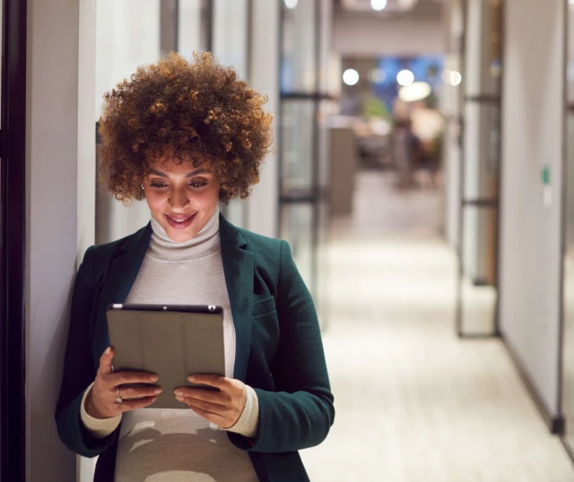 woman in the office hallway leaning against the wall with tablet in hand